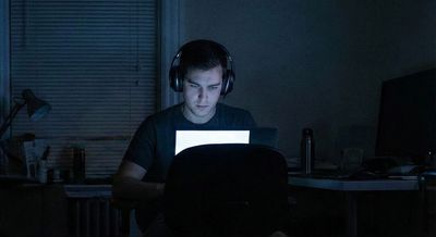 A realistic photograph of a focused cybersecurity researcher working late at night, sitting on a bed with a laptop sitting on their lap and headphones on, illuminated only by the screen in a dimly lit room.
